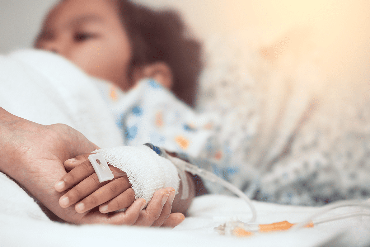 Young child laying in hospital bed, with adult's hand holding child's hand; child's hand is wrapped in bandage with cannula inserted